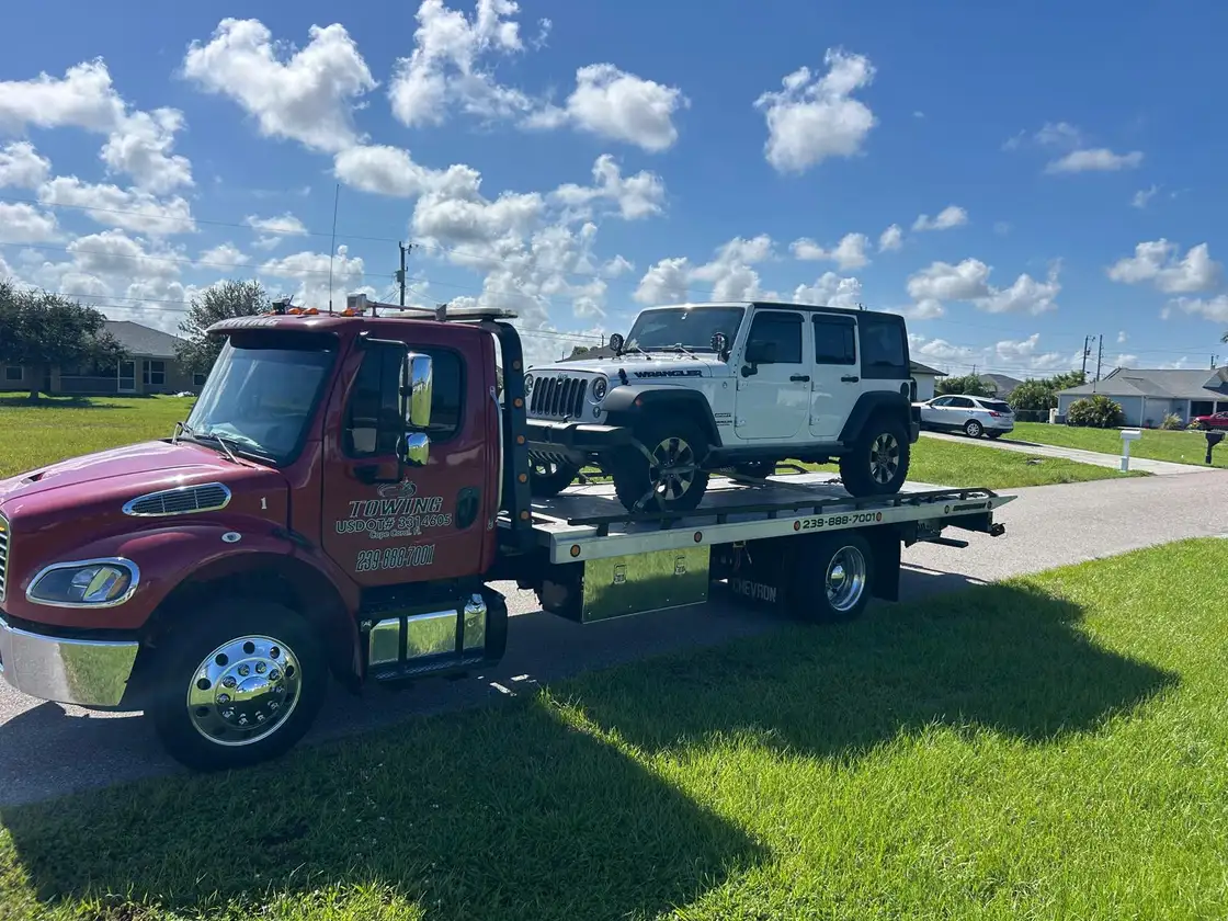 Red flatbed tow truck carrying a white Jeep Wrangler under a blue sky