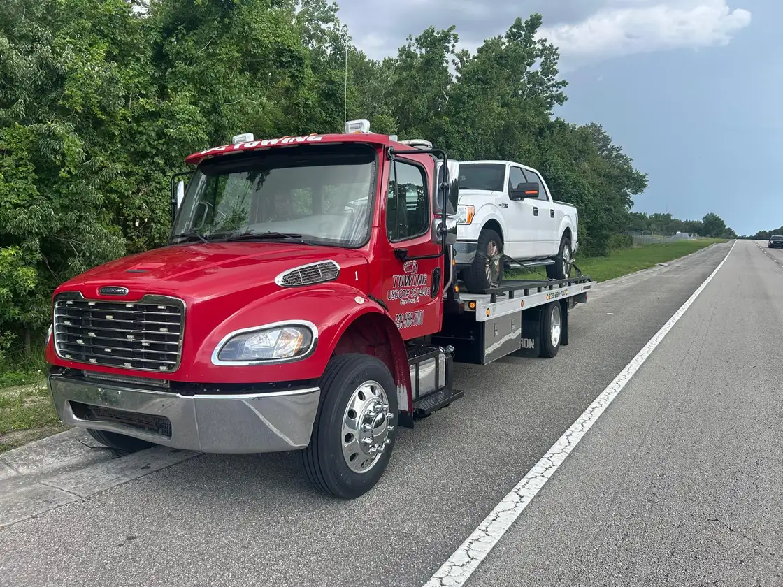La grúa de plataforma roja de Alejo’s Towing a la orilla de una carretera, con una pickup blanca encima